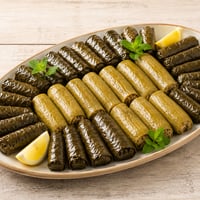 لفائف ورق عنب تحيط بصف من حبات الكوسا المحشوة الكاملة في طبق بيضاوي فاتح بحافة طينية، مع شرائح ليمون وأغصان نعناع، على طاولة خشبية مبيضة