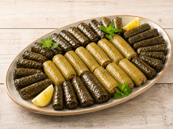 لفائف ورق عنب تحيط بصف من حبات الكوسا المحشوة الكاملة في طبق بيضاوي فاتح بحافة طينية، مع شرائح ليمون وأغصان نعناع، على طاولة خشبية مبيضة