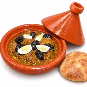 طاجين لحم بالبرقوق وأربع قطع بيض مسلوق في صحن فخاري، مع غطاء الطاجين وخبز بلدي على خلفية بيضاء